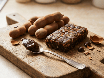 Whole brown tamarind pods, a pressed block of tamarind pulp and a spoon of dark tamarind paste on a wooden board
