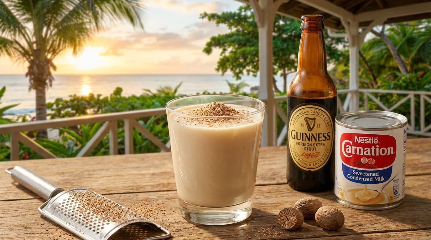 A glass of creamy, pale-brown Caribbean Guinness punch topped with grated nutmeg, beside a bottle of stout and condensed milk.
