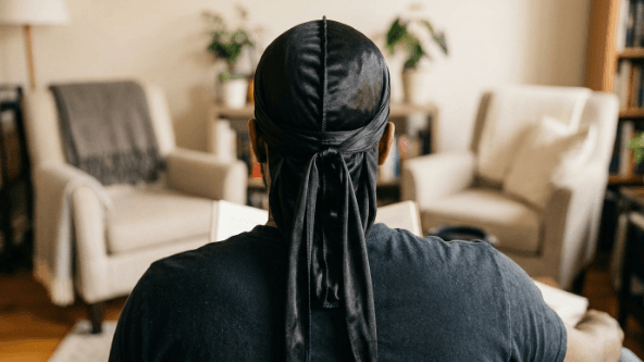 A person wearing a black silky durag tied neatly at the back, the long ties hanging down, worn to lay down and protect the hair.