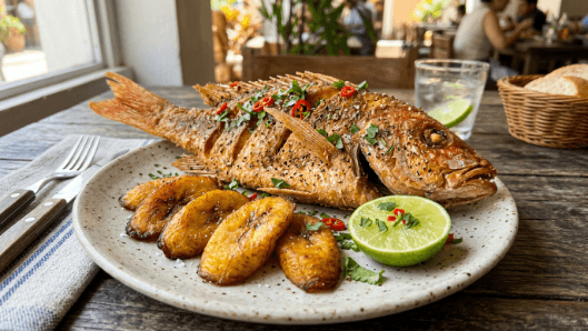 A crisp golden fried whole fish seasoned with pepper and herbs, on a plate with lime and fried plantain.