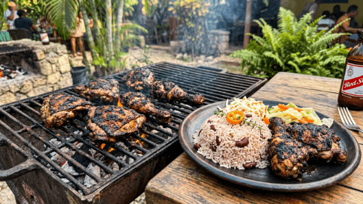 Pieces of Jamaican jerk chicken charred on a grill, dark and spiced, beside rice and peas.